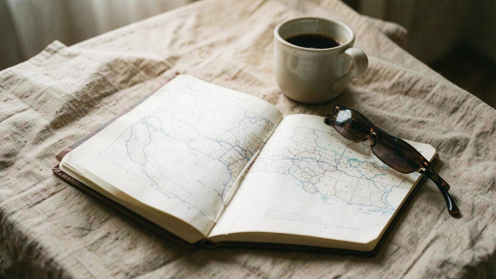 An open notebook with a printed map, a coffee cup and sunglasses on a linen table